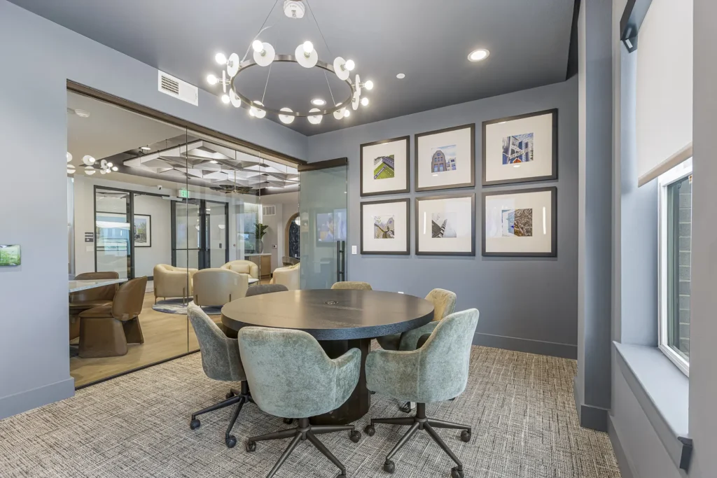 A modern conference room with a round table, four green swivel chairs, a large chandelier, framed artwork on the wall, glass doors, and a window letting in natural light.