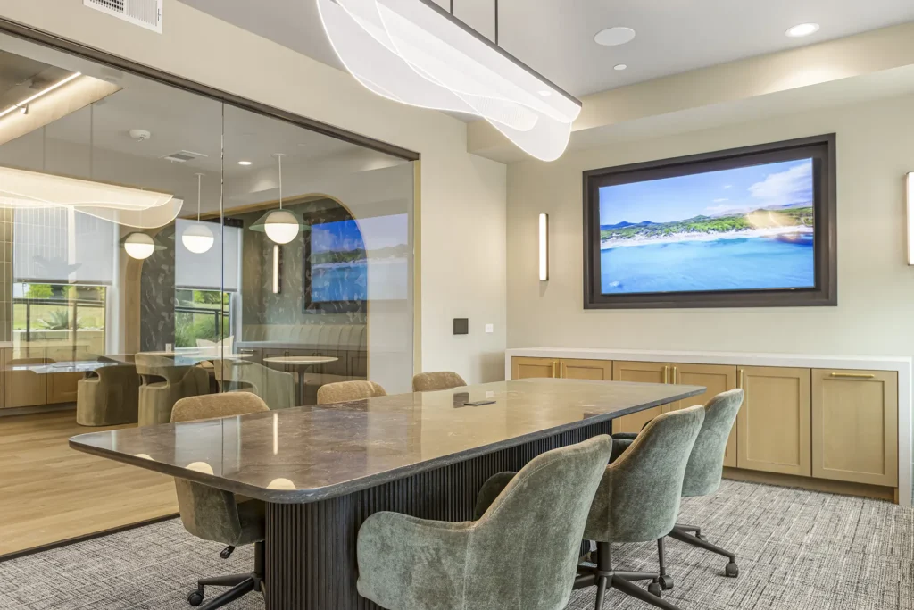 Modern conference room with a marble table, six green chairs, a large wall-mounted TV displaying a scenic landscape, and glass walls showing an adjacent meeting area with similar decor.