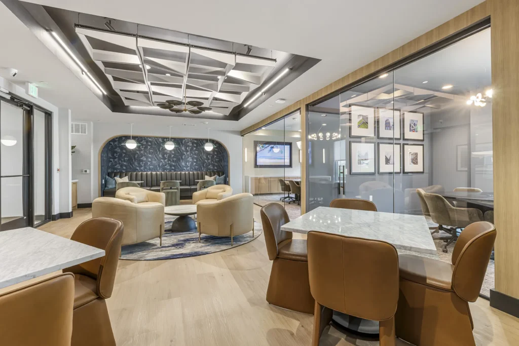 Modern lounge area with marble tables, tan leather chairs, a blue accent wall, decorative ceiling, and a glass-walled meeting room. The space is bright with contemporary furnishings and art on the walls.