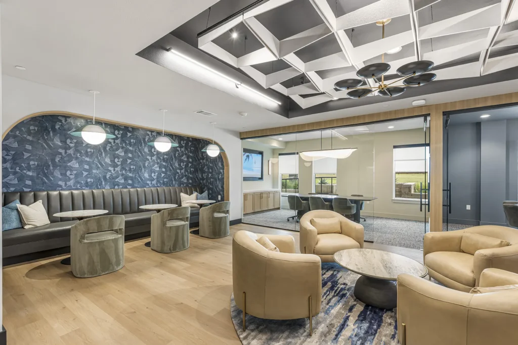 Modern office lounge with tan leather chairs, round tables, and a booth seating area with patterned blue wallpaper. There’s a glass-walled conference room in the background and geometric ceiling designs throughout.
