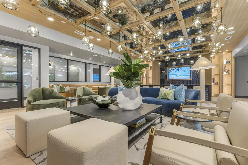 Modern lounge with plush seating, a large plant centerpiece on a coffee table, stylish ceiling lights, and a TV in the background. The room features neutral and blue tones with decorative accents.
