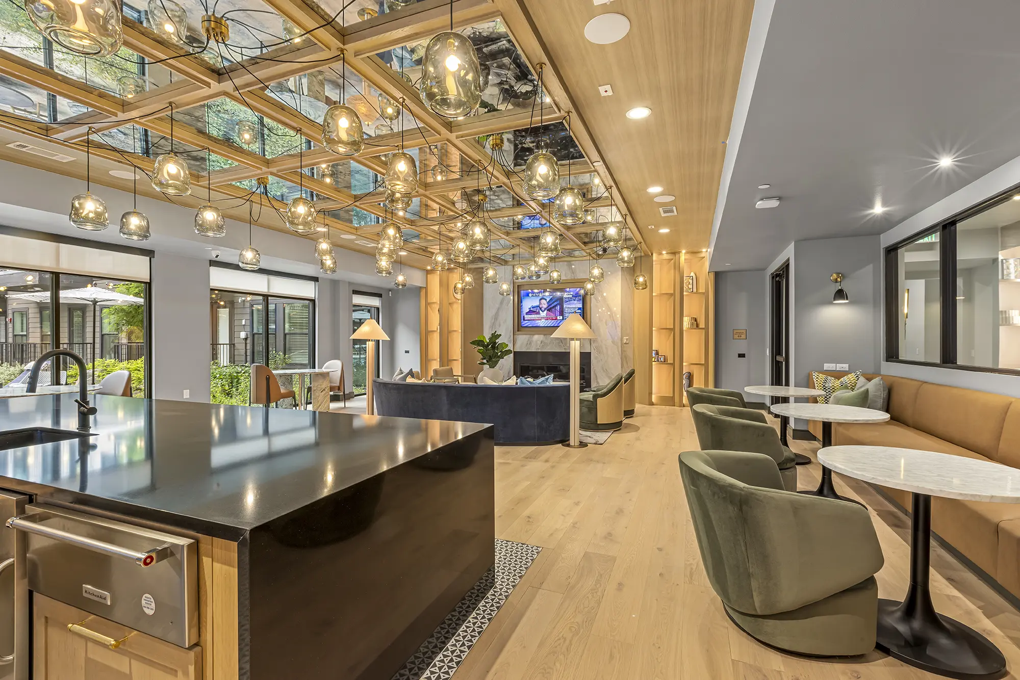 Modern lounge area with stylish light fixtures, mirrored ceiling panels, green chairs around small tables, a large kitchen island, and a TV above a fireplace, with large windows letting in natural light.