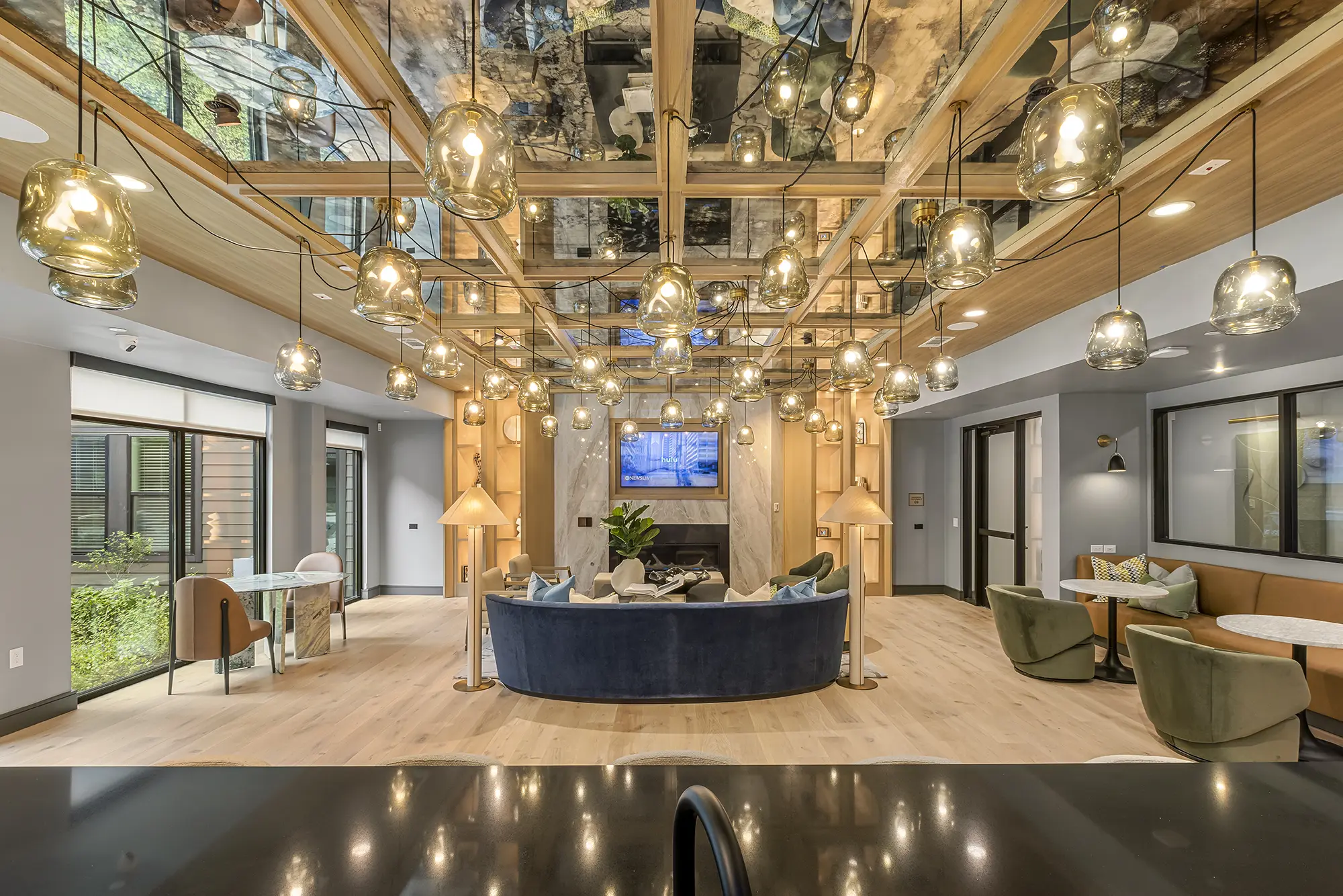 Elegant lounge with modern decor, featuring a blue curved sofa, round tables, armchairs, wooden floors, and numerous hanging glass pendant lights. A TV is mounted above a fireplace, and large windows bring in natural light.