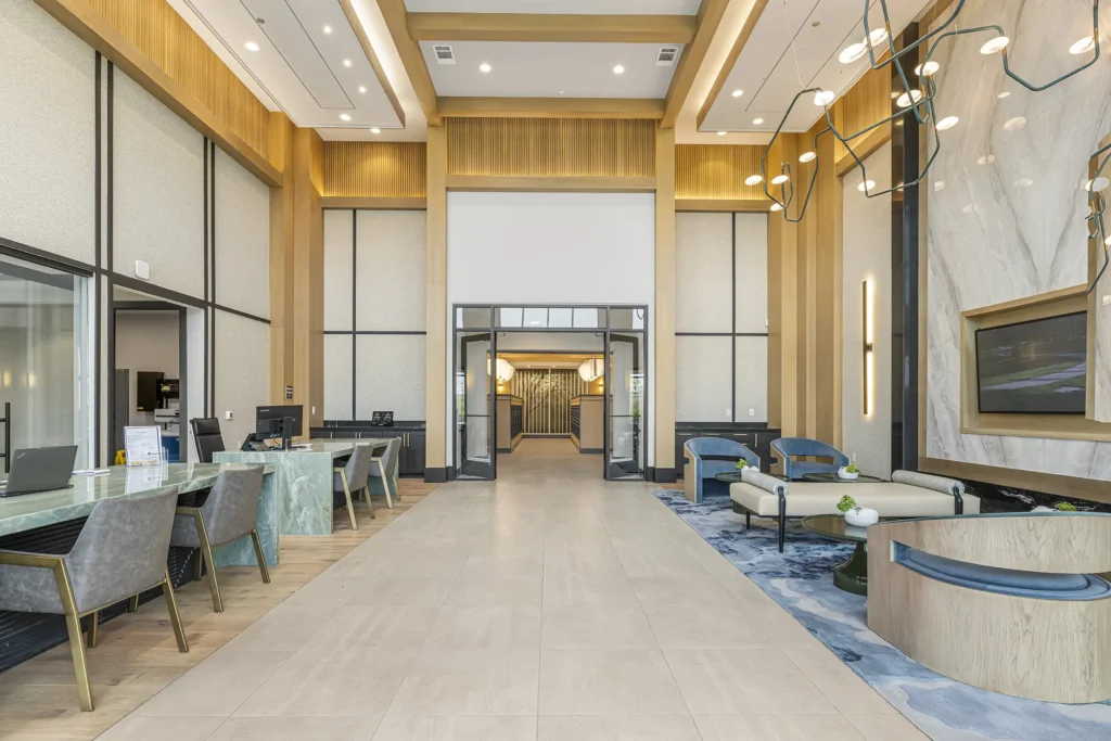 Modern hotel lobby with high ceilings, elegant light fixtures, marble and wood accents, a seating area with sofas and chairs, desks with chairs on the left, and a large decorative wall on the right.