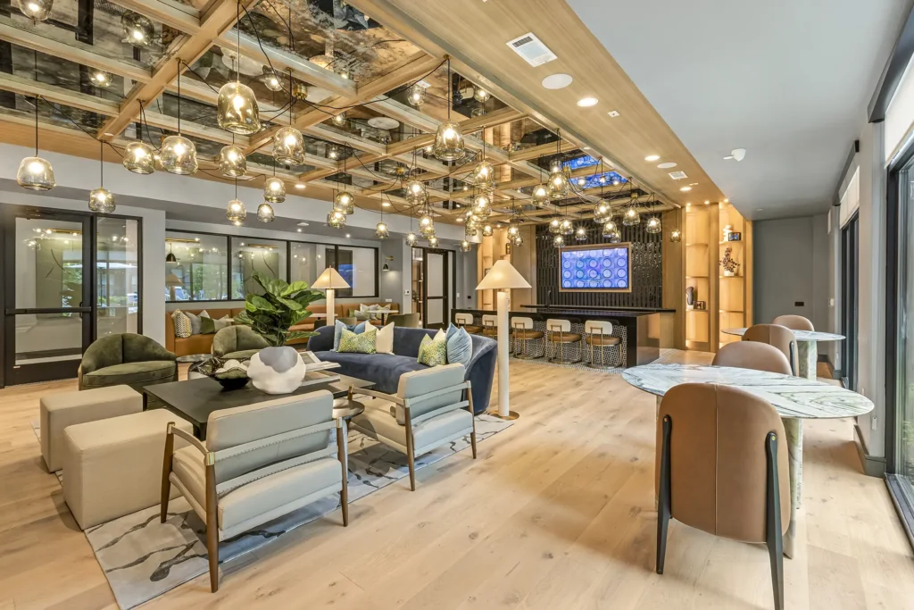 A modern lounge with cozy seating, a bar area, and large windows. Pendant lights hang from a wooden ceiling grid, reflecting in mirrors above. The space features neutral tones, plants, and a wall-mounted TV.