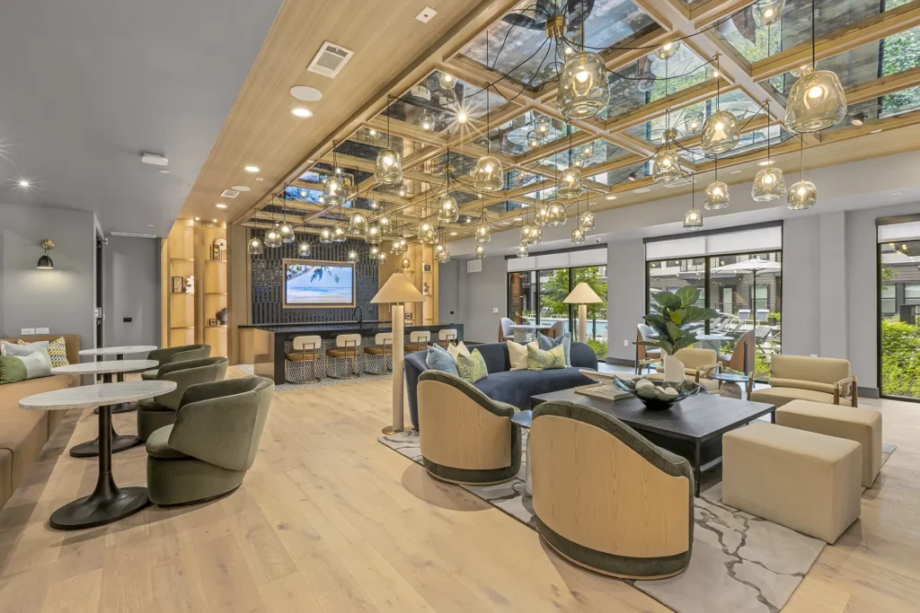 Modern lounge with wood floors, green and beige chairs, sofas, marble tables, and a TV. The ceiling has mirrors and hanging lights. Large windows let in natural light and show greenery outside. The setting looks stylish and inviting.