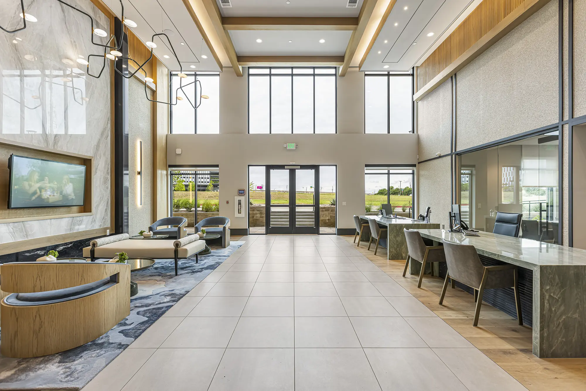 Modern, spacious lobby with high ceilings, large windows letting in natural light, stylish light fixtures, seating area with chairs and sofas, and desk areas along the right wall. Neutral tones and contemporary design elements.