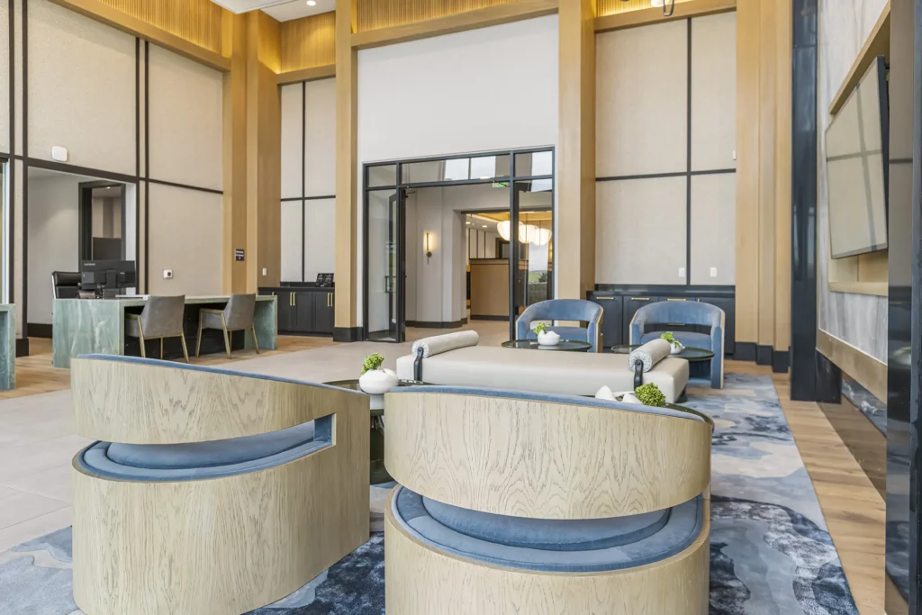 Modern lounge area with light wood and blue furniture, round chairs, and a large coffee table. Tall ceilings, large windows, and elegant decor create an open, inviting atmosphere.