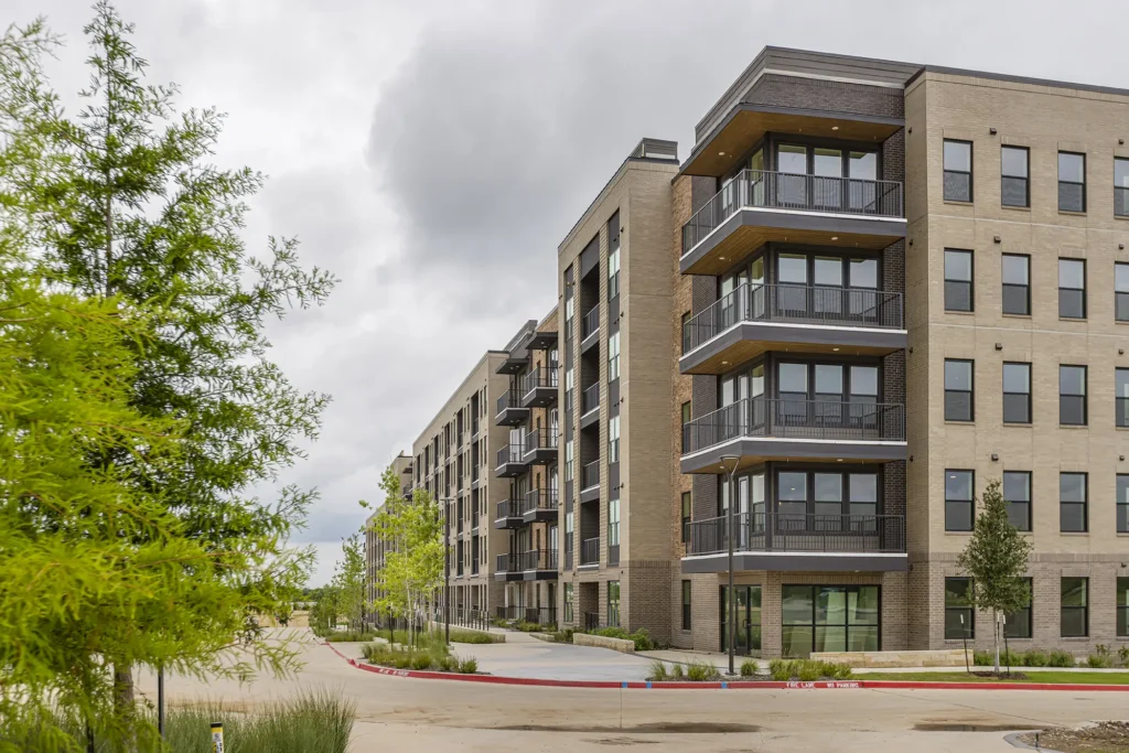 Modern multi-story apartment building with balconies, large windows, and brick exterior, set along a curved, landscaped street with young trees and cloudy skies overhead.