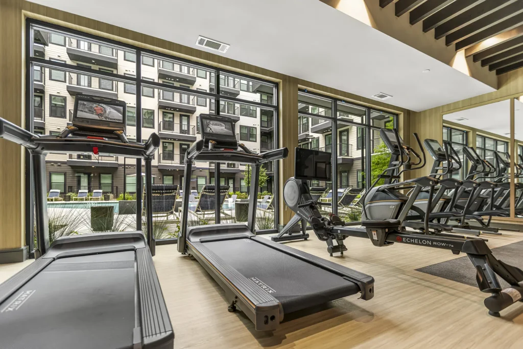 Modern gym with treadmills, stationary bikes, and rowing machines facing large floor-to-ceiling windows. Outside, an apartment complex and pool area are visible through the glass.