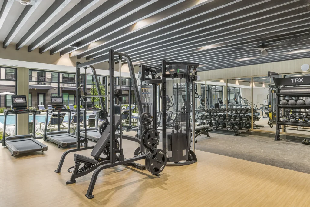 A modern gym with treadmills, weight machines, free weights, and large mirrors. Windows on one side reveal an outdoor pool area. The space has light wood floors and exposed ceiling beams.