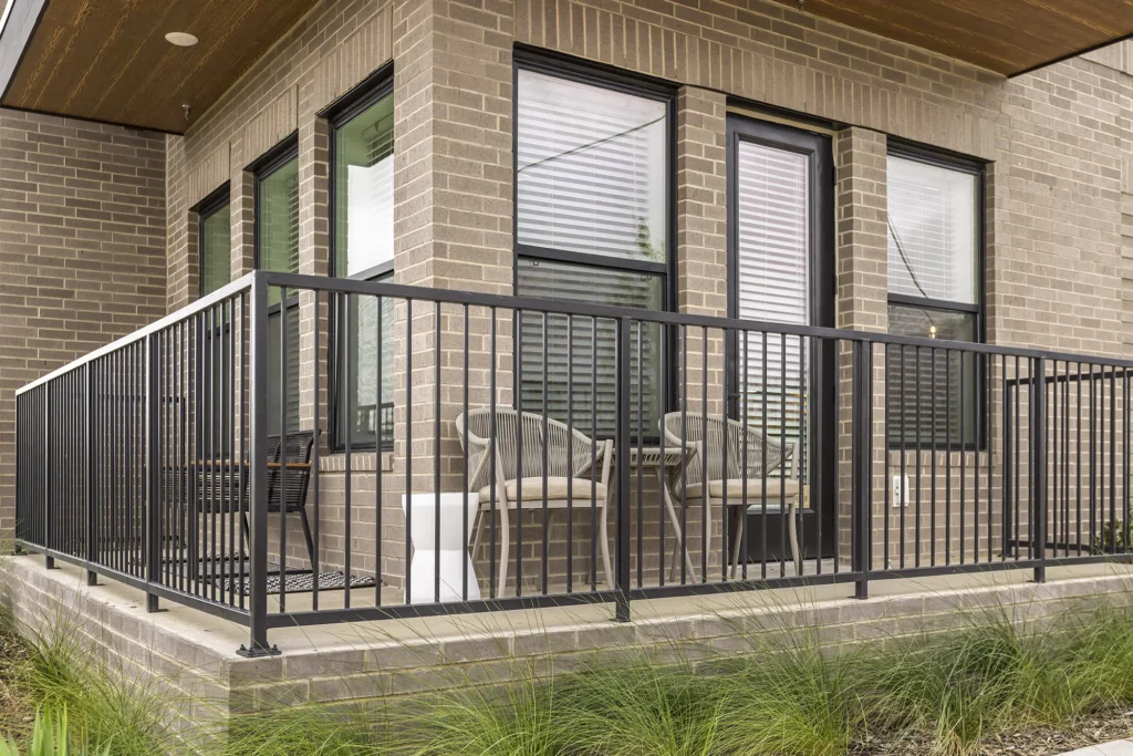 A modern brick patio with black metal railing, two woven chairs, a small side table, and large windows with white blinds on a corner of a building. Green ornamental grass borders the base of the patio.