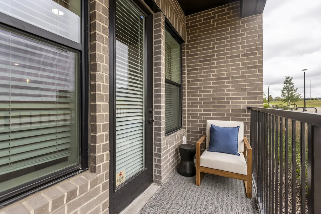 A small patio with a cushioned wooden chair, blue pillow, black side table, and black metal railing. The patio is adjacent to a brick wall and a glass door with blinds, overlooking a grassy area.