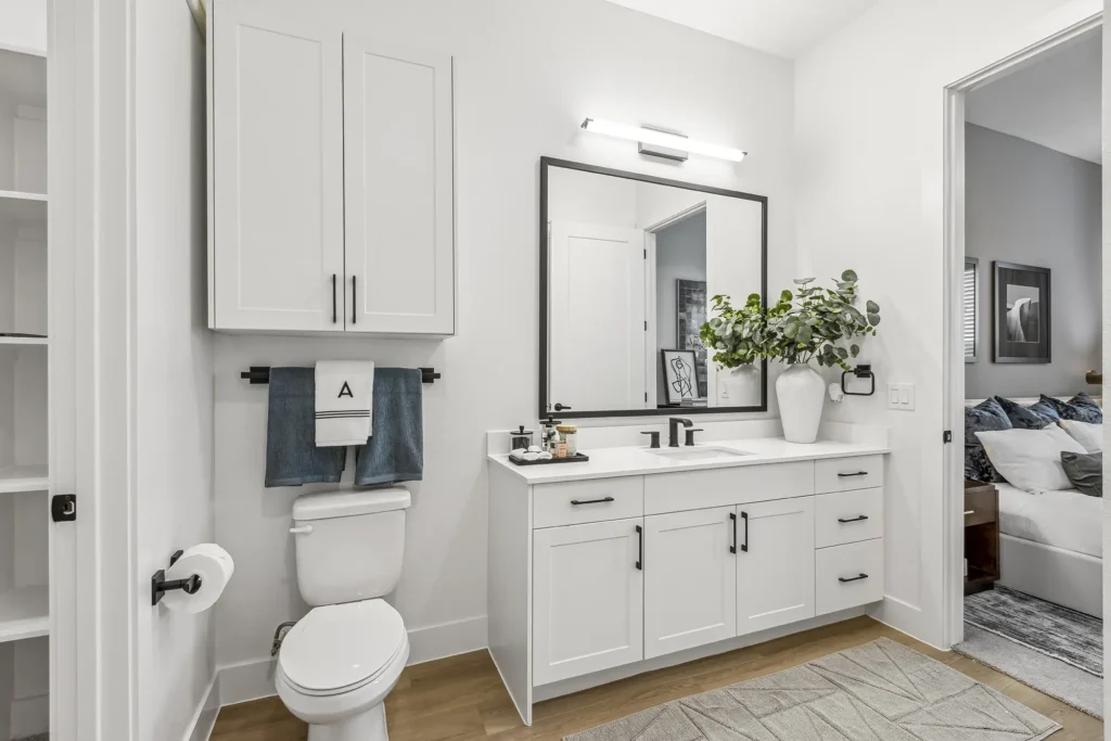 Modern white bathroom with a double-sink vanity, large mirror, potted plant, and cabinets. A toilet with a towel rack is beside the vanity. The floor is wood, and a bedroom is visible through an open door.