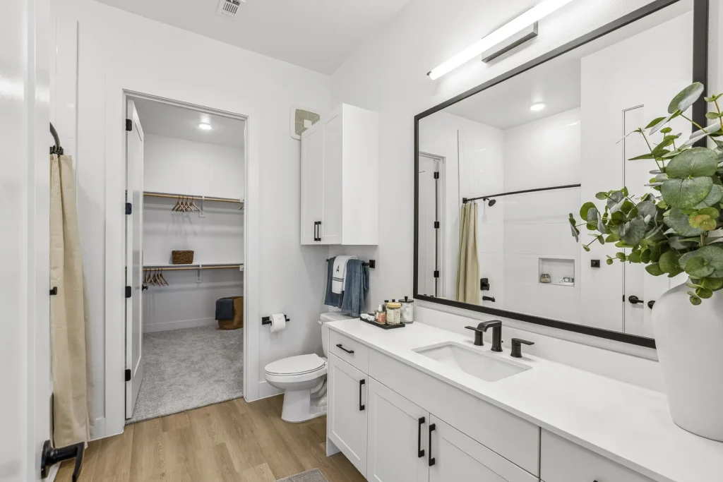 Modern bathroom with white walls, a double-sink vanity, large mirror, black fixtures, and wood-look flooring. A toilet and towels are beside the vanity. An open doorway leads to a spacious walk-in closet with carpet.