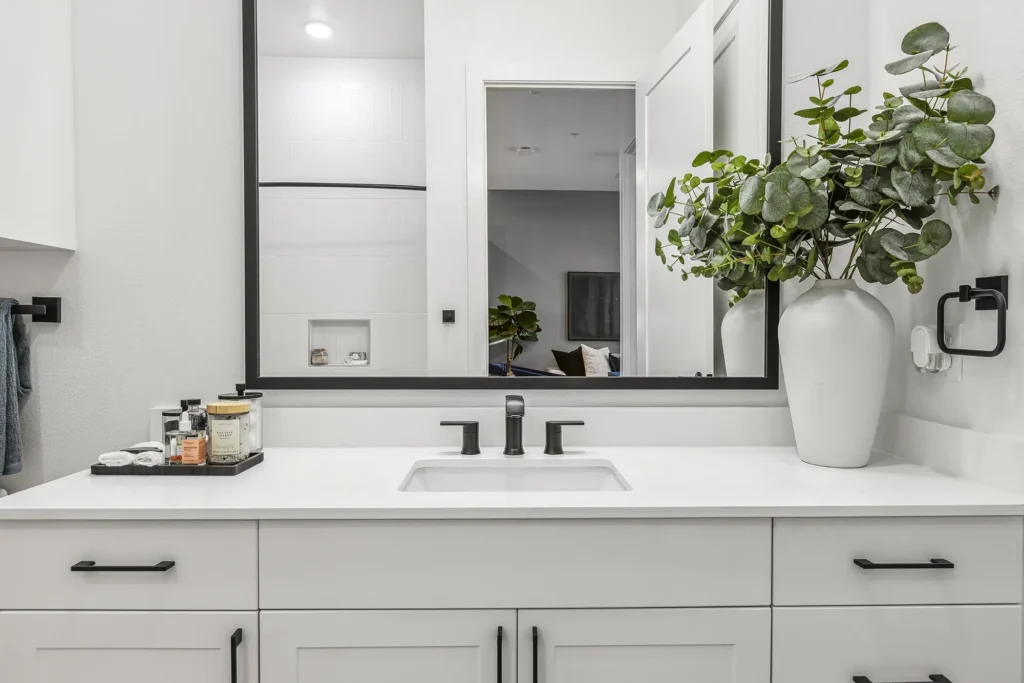 Modern bathroom vanity with a white countertop, black fixtures, a large mirror, a white vase with green foliage, and various toiletries neatly arranged on a tray beside the sink.