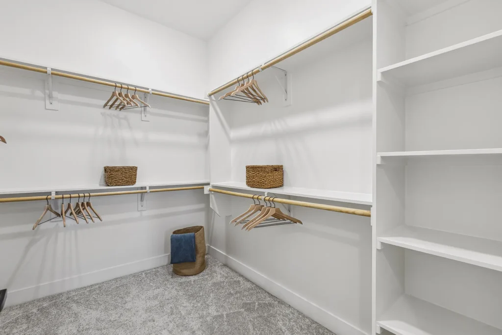 A spacious walk-in closet with white shelves, wooden hanging rods, empty wooden hangers, woven baskets, and a fabric laundry hamper on a light carpeted floor. The walls are white and the space is neatly organized.