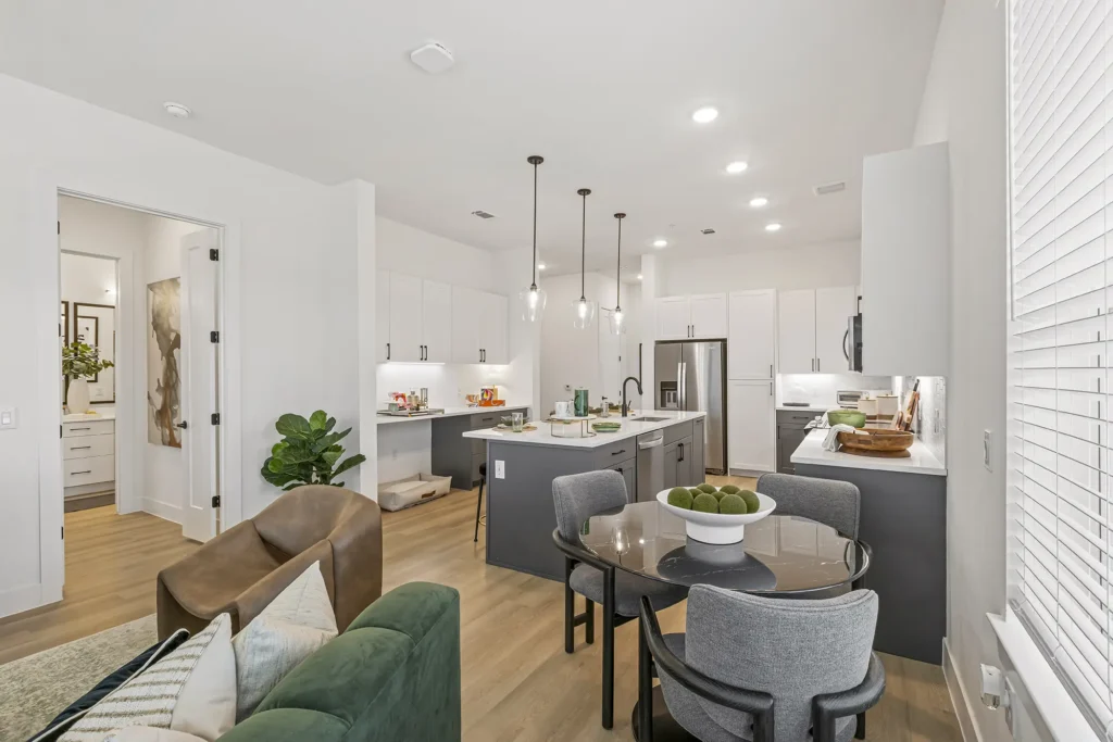 Modern open-concept kitchen and dining area with white cabinets, stainless steel appliances, a round dining table with chairs, pendant lights, and a living space with a green sofa and armchair.