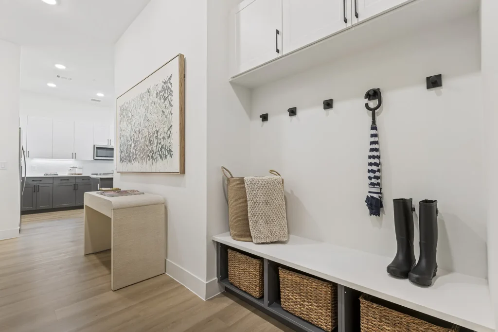 A modern mudroom with hooks on the wall, a striped scarf, black rain boots, woven baskets below a bench, and two wicker baskets with a blanket. The space connects to a kitchen with white cabinetry.