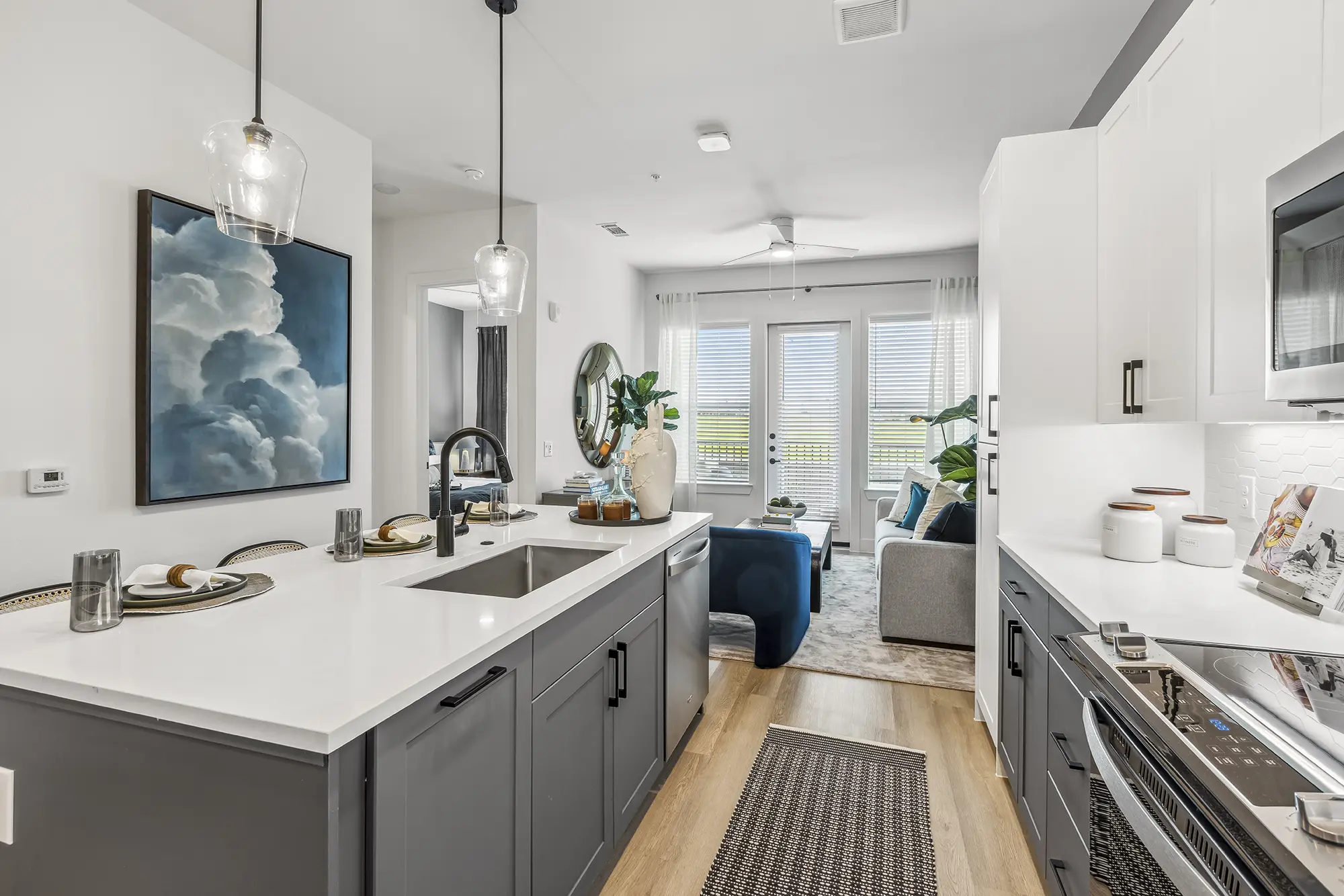 Modern kitchen with gray and white cabinets, a center island with sink and bar seating, pendant lights, and a view into a bright living room with a sofa, plants, and large windows with blinds.