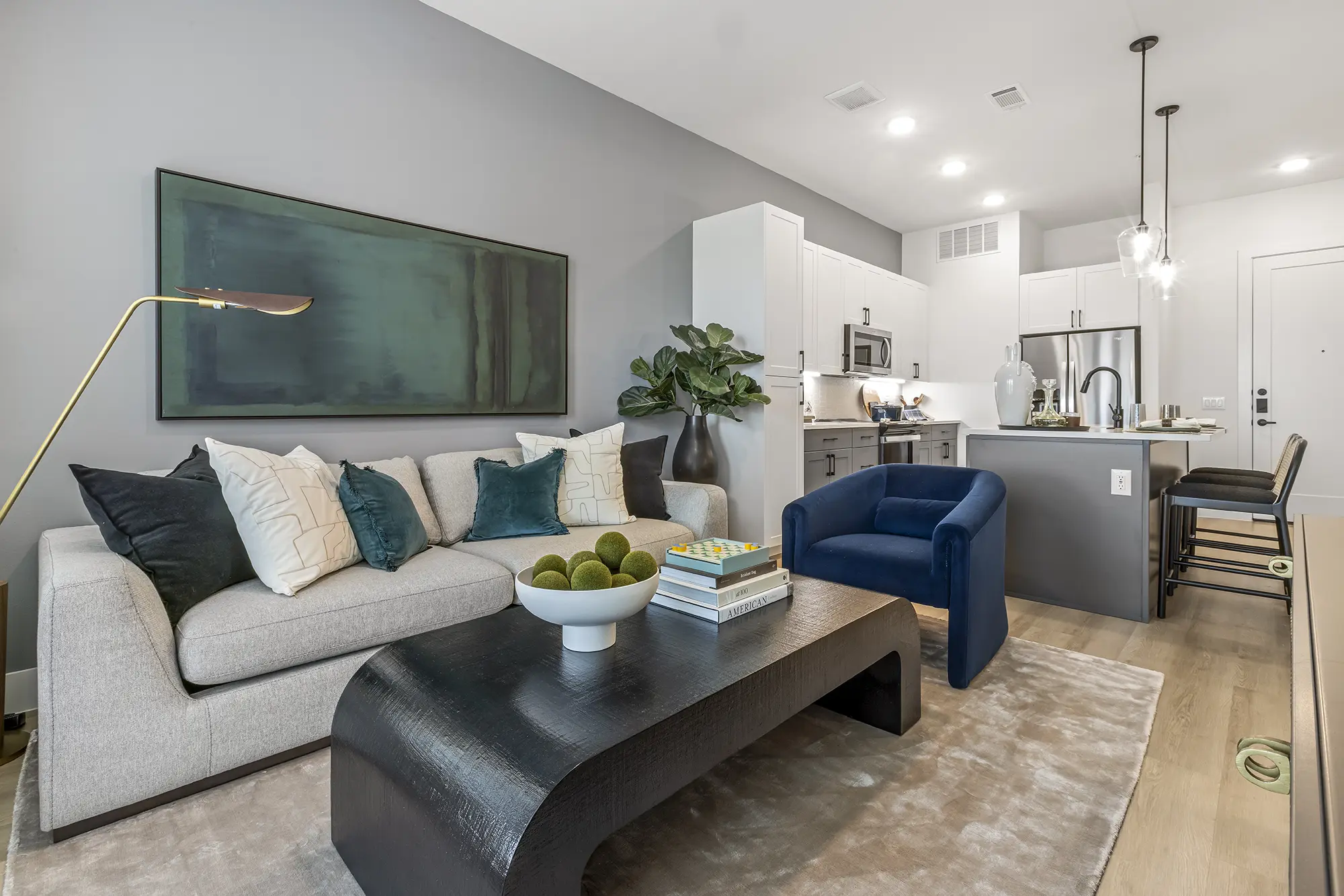 Modern living room with a light gray sofa, assorted pillows, blue armchair, black coffee table with decor, open kitchen in the background, and large abstract wall art above the sofa. Light wood floors and pendant lights complete the space.