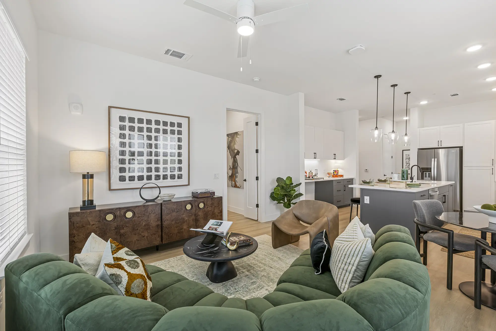 Modern living room with a green velvet sectional sofa, round black coffee table, decorative pillows, and a wooden sideboard. The space flows into a kitchen with white cabinets, island, and pendant lights.