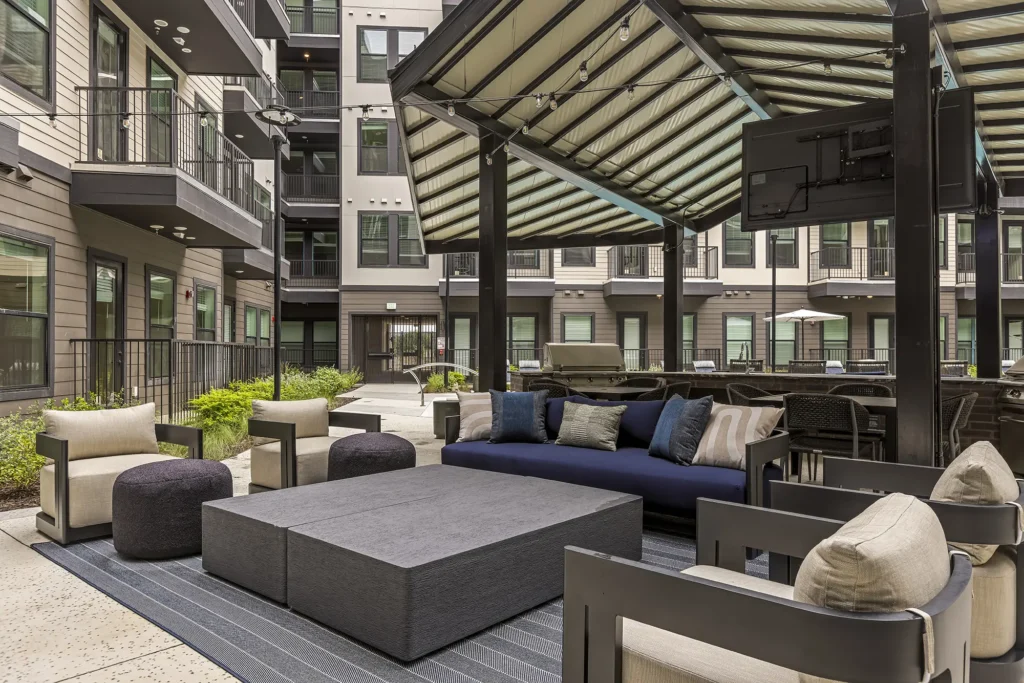 Modern outdoor patio with cushioned seating, armchairs, and low tables under a large metal pergola in a residential apartment courtyard. Surrounding buildings have balconies and large windows.
